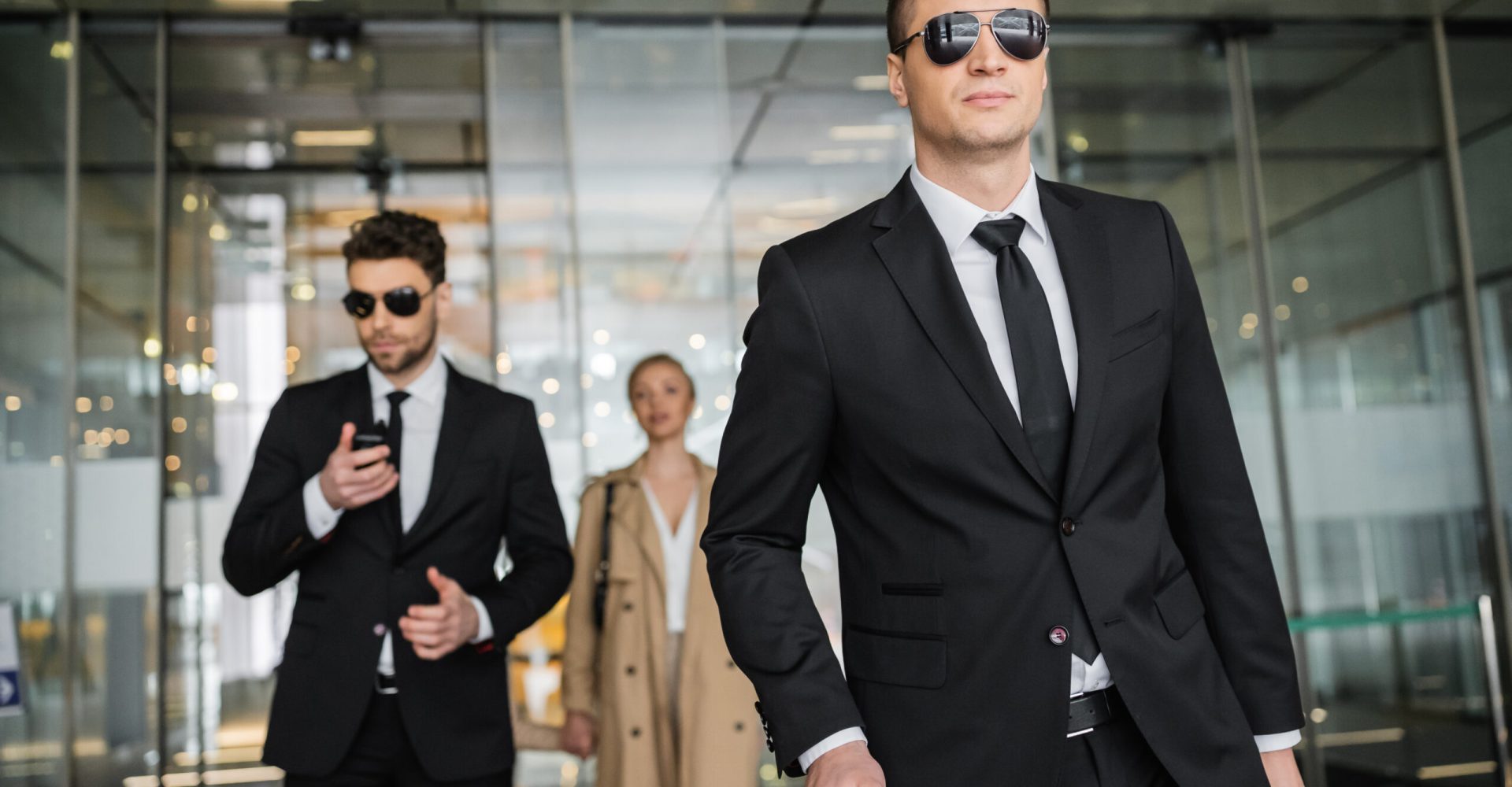 private security service, personal safety concept, handsome bodyguards in formal wear and sunglasses protecting safety of blonde client on blurred background, men and woman walking in hotel private security service, personal safety concept, handsome bodyguards in formal wear and sunglasses protecting safety of blonde client on blurred background, men and woman walking in hotel